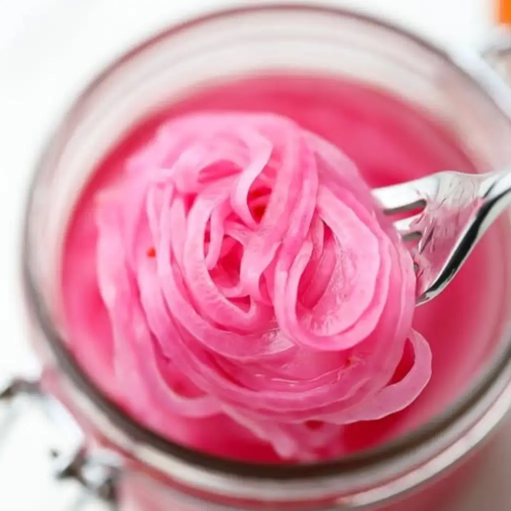 A close-up image of pickled red onions being lifted with a fork from a mason jar, showcasing the vibrant pink brine and thin onion slices.