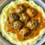 Meatballs topped with savory gravy served on a bed of creamy mashed potatoes, garnished with fresh parsley for a comforting meal.