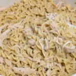 A close-up image of Crockpot Chicken and Noodles, showing creamy broth, tender chicken, and soft egg noodles stirred with a wooden spoon.