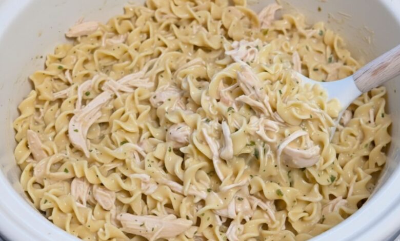 A close-up image of Crockpot Chicken and Noodles, showing creamy broth, tender chicken, and soft egg noodles stirred with a wooden spoon.