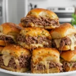 A plate of freshly made French Dip Sliders stacked high, with tender roast beef, melted cheese, and golden, soft slider buns topped with herbs.