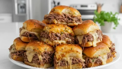 A plate of freshly made French Dip Sliders stacked high, with tender roast beef, melted cheese, and golden, soft slider buns topped with herbs.