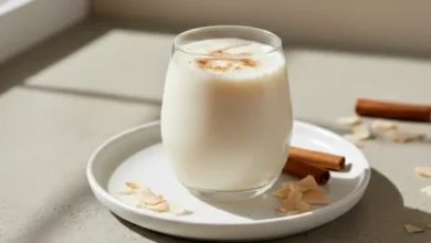 Creamy homemade coquito served in a glass, topped with cinnamon, on a white plate with coconut flakes and cinnamon sticks.