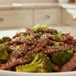 Beef and broccoli stir-fry with sesame seeds and green onions on a white plate.