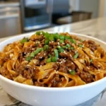 Bowl of Mongolian noodles with ground beef, garnished with green onions and sesame seeds, served on a kitchen countertop.