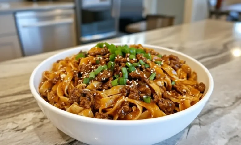 Bowl of Mongolian noodles with ground beef, garnished with green onions and sesame seeds, served on a kitchen countertop.