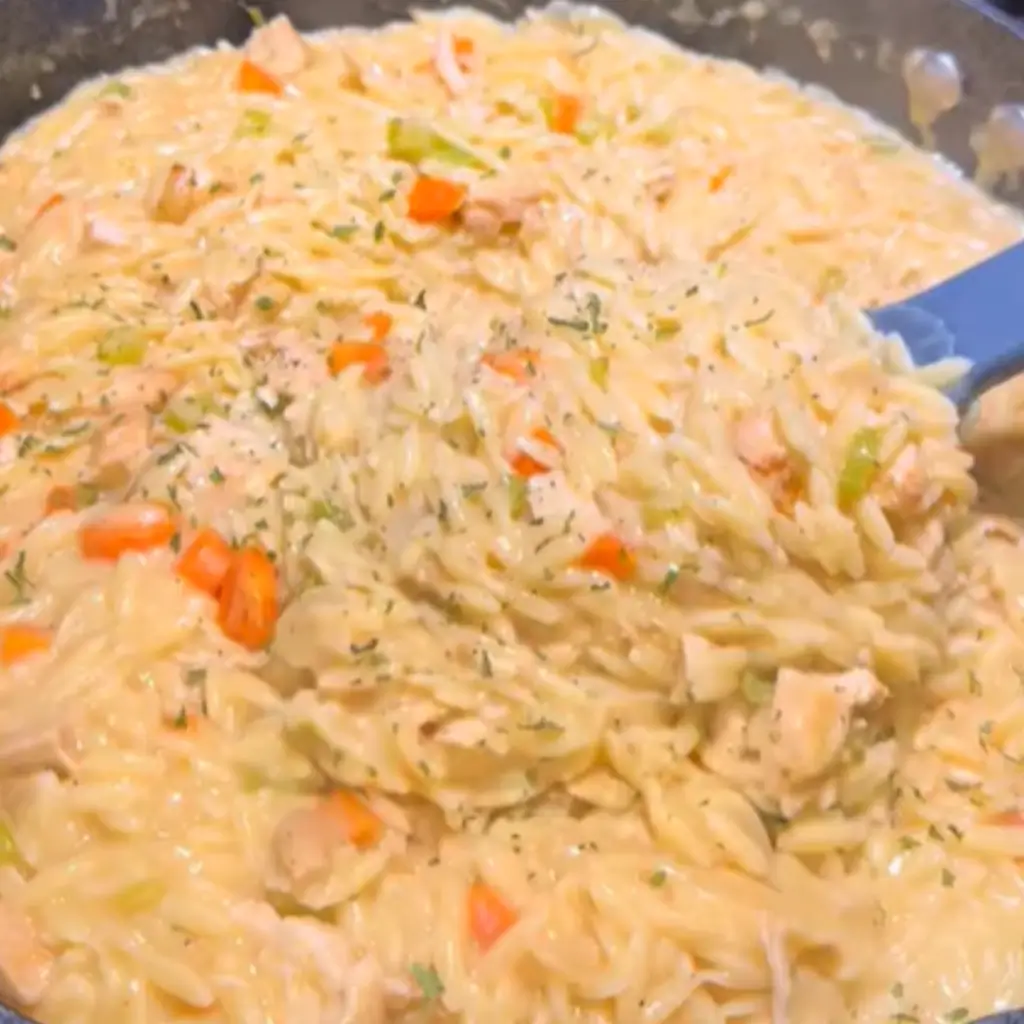 Close-up of creamy chicken pot pie orzo in a skillet, with chunks of chicken, carrots, and green vegetables being stirred with a spatula.