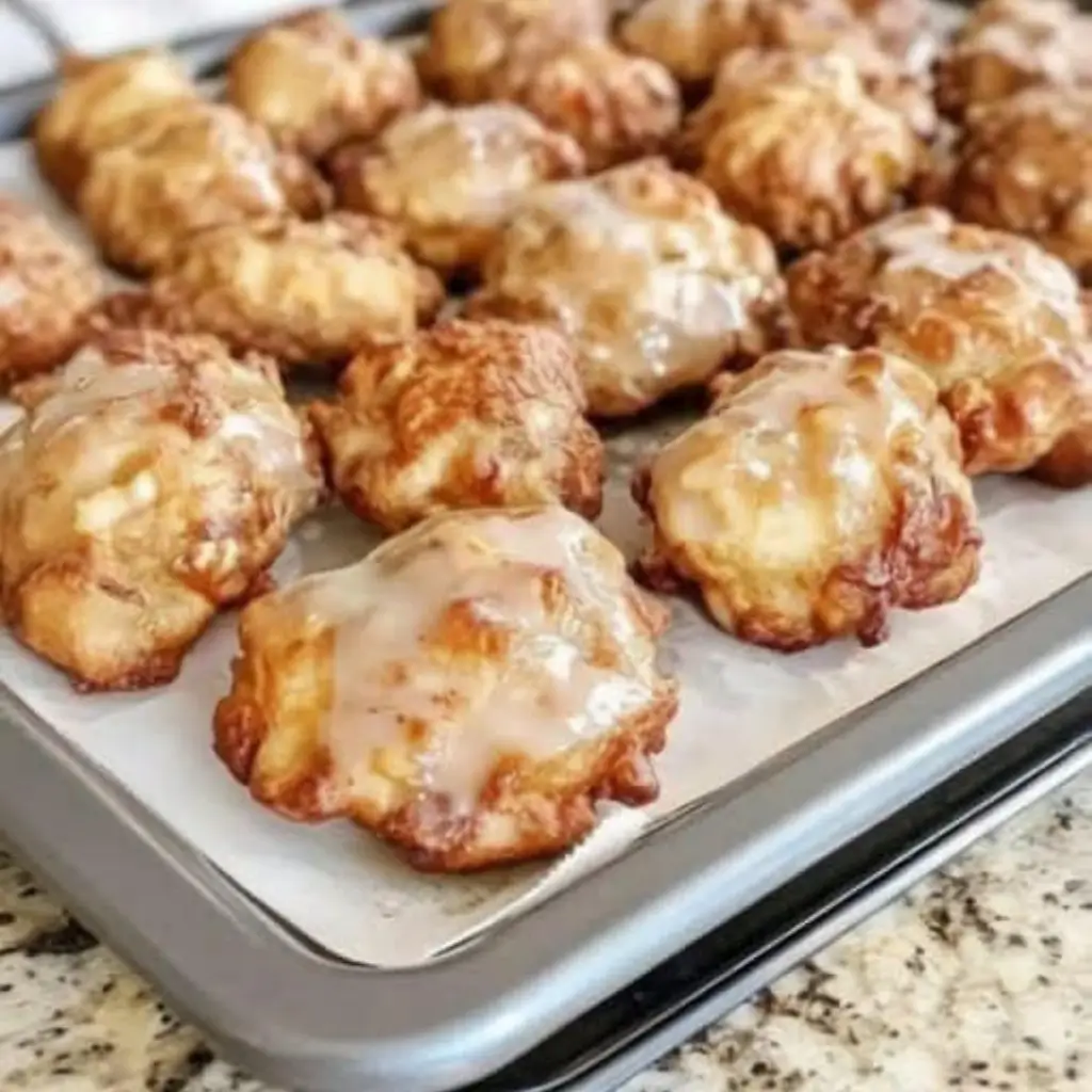 A tray of freshly baked apple fritters with a golden-brown, crispy exterior and a sweet glaze drizzled on top, arranged neatly on parchment paper.