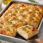 Freshly baked sourdough focaccia with a golden-brown crust, topped with rosemary, sea salt, and garlic, resting in a metal tray with olive oil and sea salt nearby.